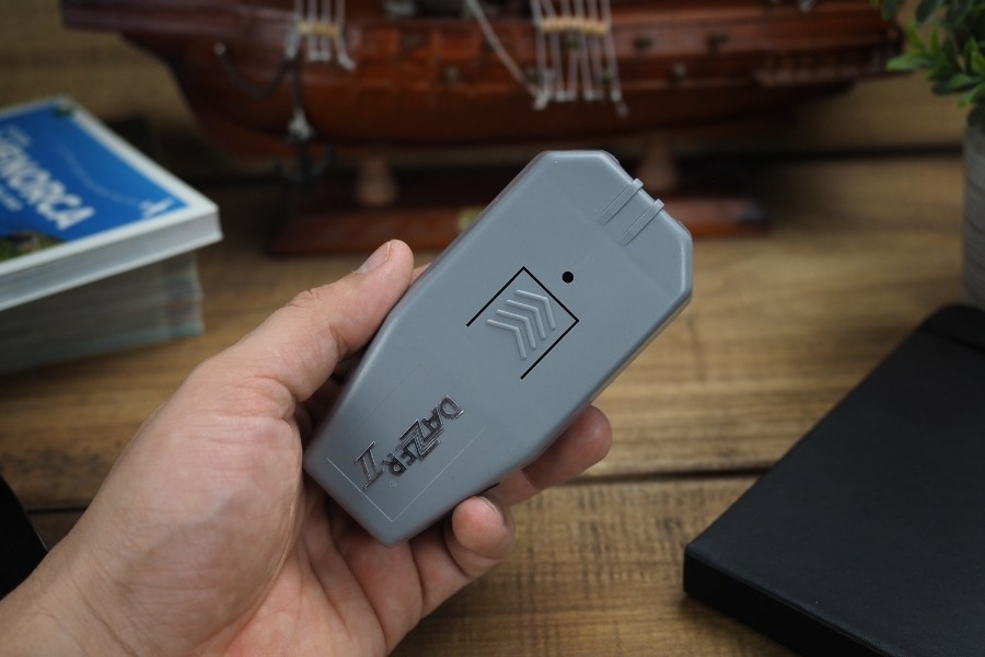A person holds a grey ultrasonic Dazer II device used to scare away dogs. In the background, books and a model ship on a wooden table.